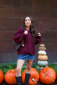 Woman in a purple sweater and blue skirt standing next to pumpkins against a wooden wall.