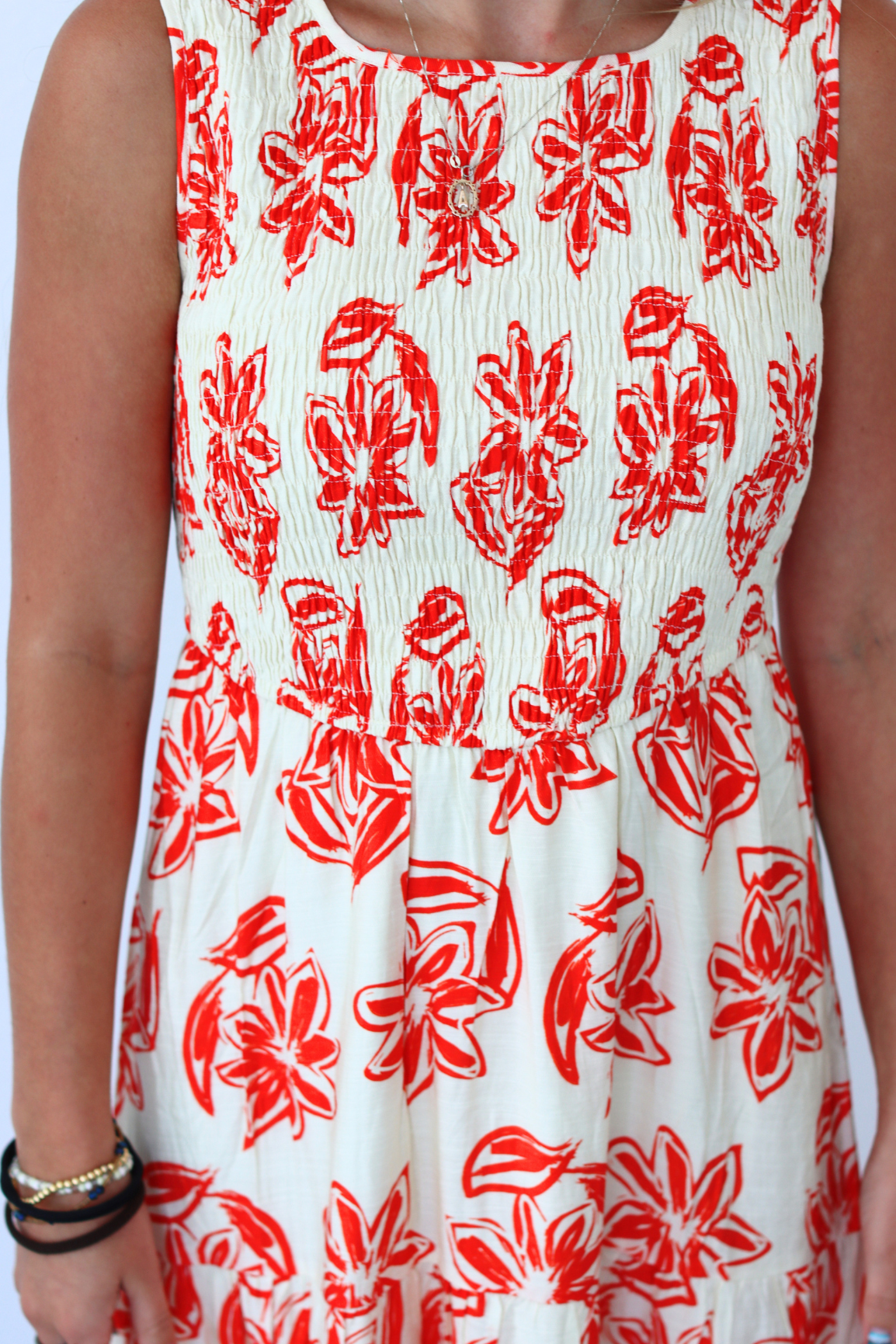 A sleeveless white dress with a red floral print, worn by a model. The dress features a smocked bodice and a gathered skirt, with a small gold neckline.