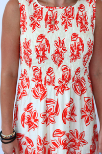 A sleeveless white dress with a red floral print, worn by a model. The dress features a smocked bodice and a gathered skirt, with a small gold neckline.