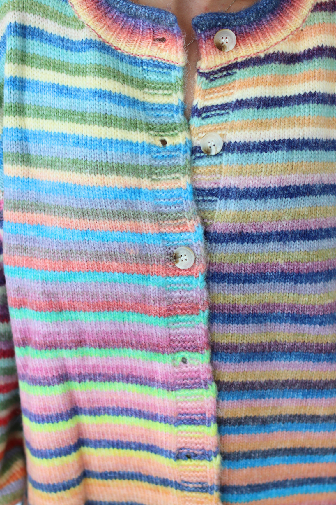 Close-up of a hand-knit long-sleeve henley shirt with horizontal rainbow stripes in pink, blue, green, yellow, orange, and purple, featuring a round collar and white buttons.