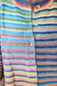 Close-up of a hand-knit long-sleeve henley shirt with horizontal rainbow stripes in pink, blue, green, yellow, orange, and purple, featuring a round collar and white buttons.