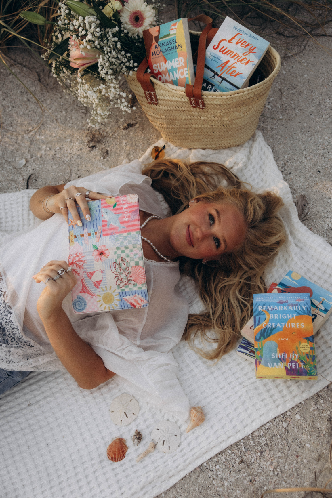 A woman lies on a white blanket outdoors, holding a colorful kindle case. A woven basket filled with books sits nearby on sand, along with flowers and a few more books scattered on the blanket.