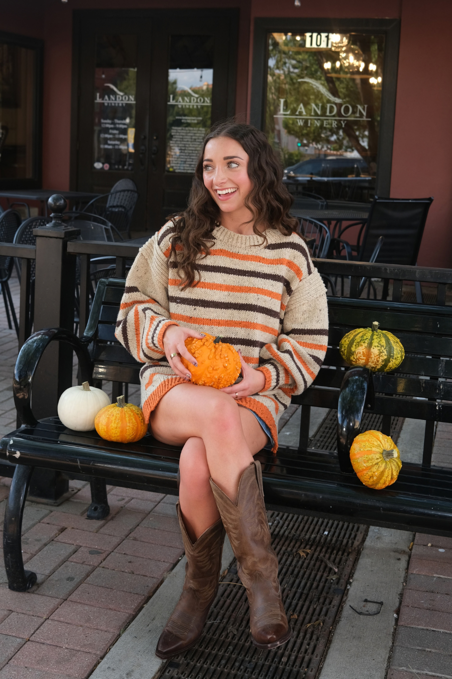 A woman wearing a chunky knit sweater with horizontal stripes in cream, orange, and dark brown, paired with a blue denim skirt