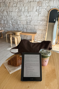 A Kindle e-reader device displaying a book, set on a wooden table in a cafe with a croissant, a brown bag, and a pink drink nearby, against a stone wall background.