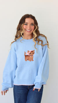 Woman wearing a light blue sweater with a cat embroidery design against a white background 