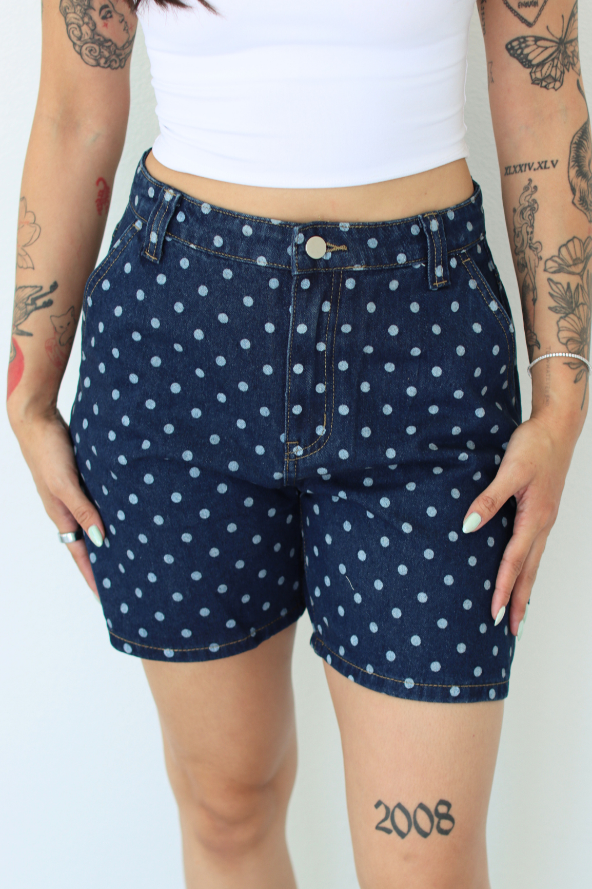 A person wearing dark blue denim shorts with a white polka dot pattern, paired with a white cropped tank top. The photo is cropped from the chest down against a plain white background.
