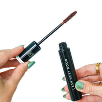 Mascara wand and tube held by a hand with green nail polish on a white background