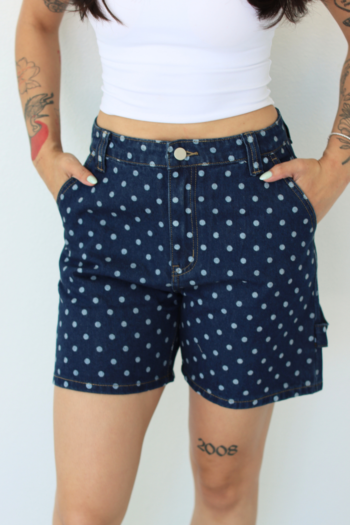 A person wearing dark blue denim shorts with a white polka dot pattern, paired with a white cropped tank top. The photo is cropped from the chest down against a plain white background.