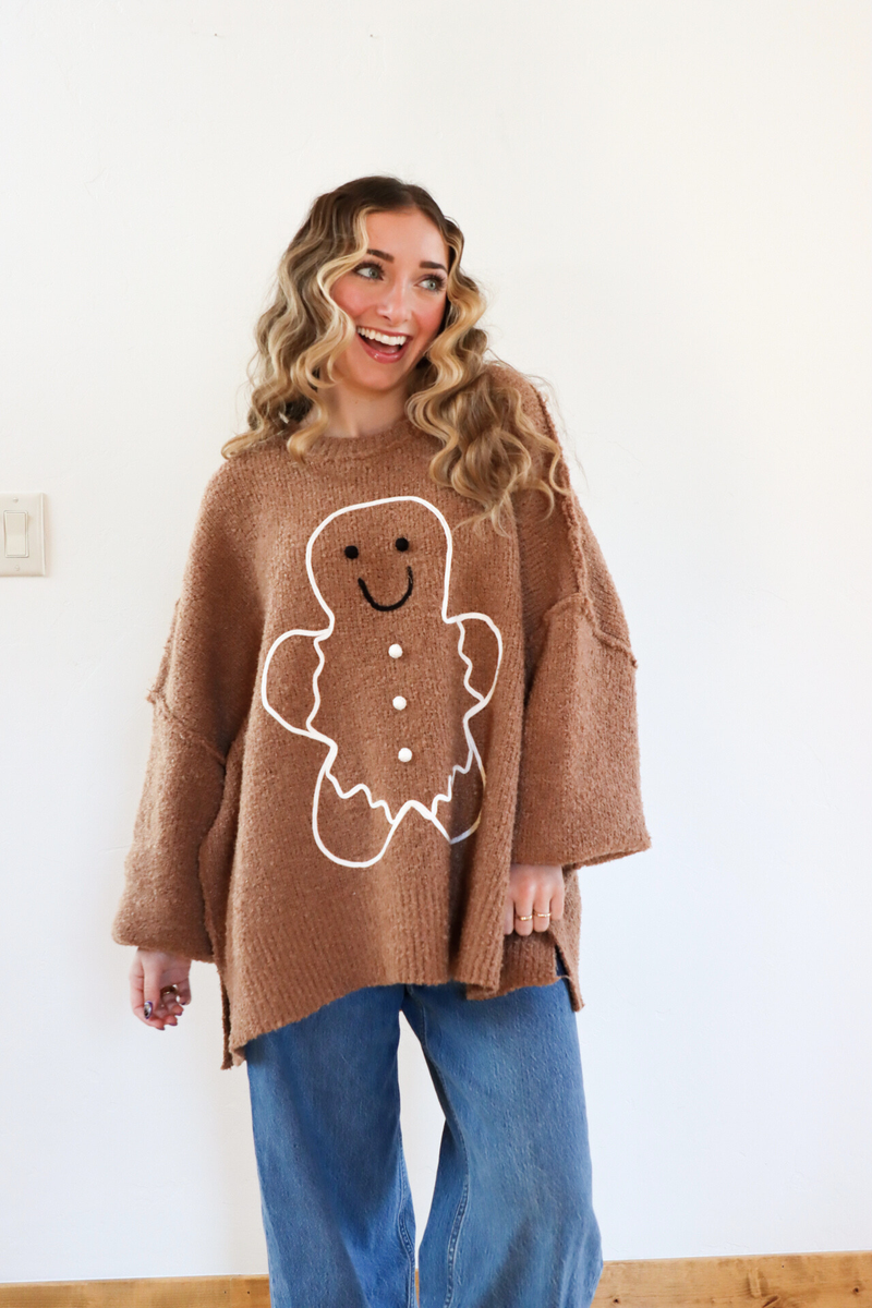 Woman wearing a brown sweater with a gingerbread man design, standing against a white wall.
