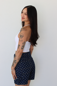 A woman with long black hair and visible tattoos is shown from the side/back wearing a white ribbed tank top and navy blue shorts with small white polka dots, standing against a plain white wall.