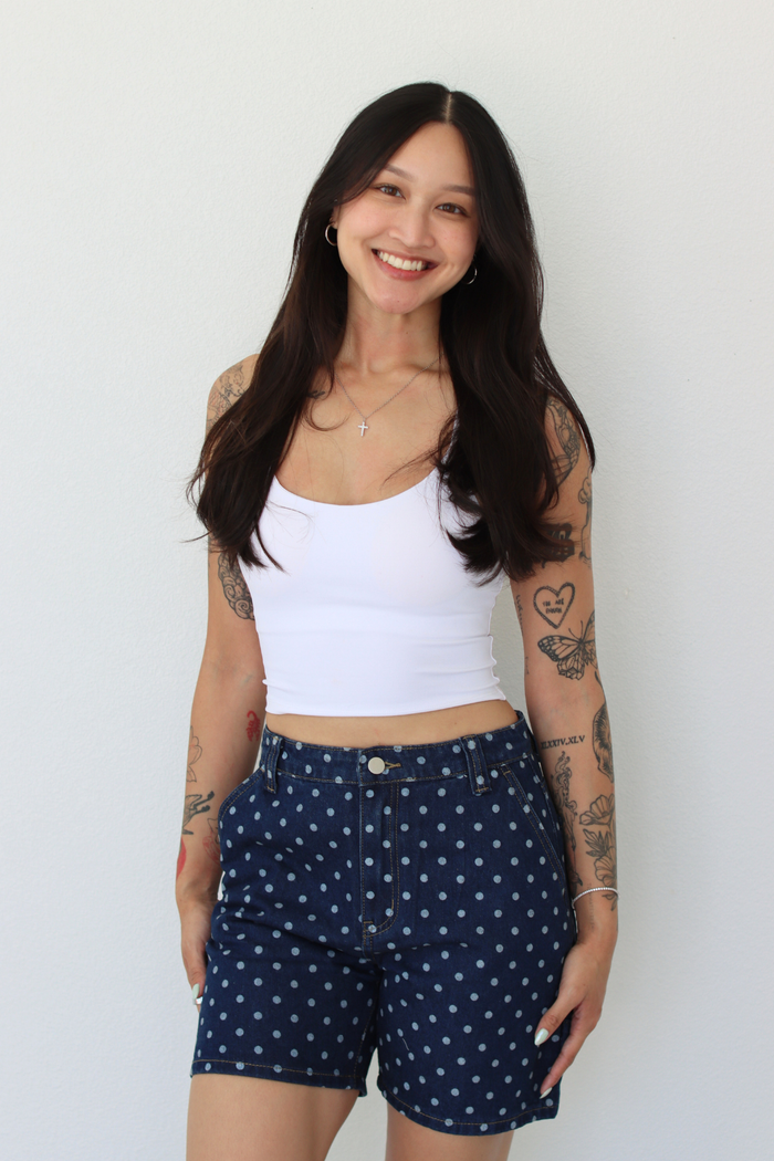 A smiling woman with long dark hair and visible arm tattoos is wearing a white ribbed tank top and navy blue shorts with a small white polka dot pattern, standing against a plain white wall.