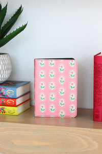Pink kindle with floral pattern on a wooden surface with books and a plant.