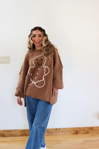 Woman wearing a brown sweater with a gingerbread man design against a white wall.