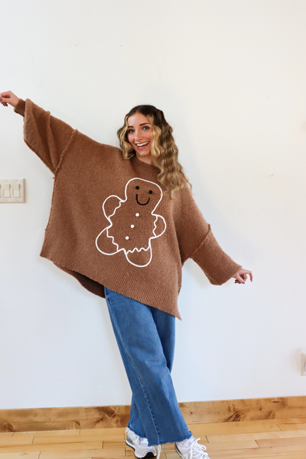 Woman wearing a brown sweater with a gingerbread man design against a white wall.