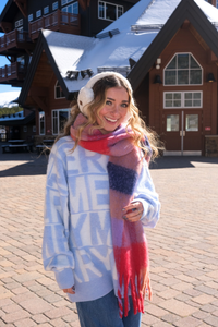 Woman wearing a colorful scarf and winter clothing in front of a wooden building.