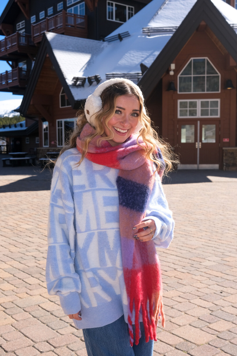 Woman wearing a colorful scarf and winter clothing in front of a wooden building.