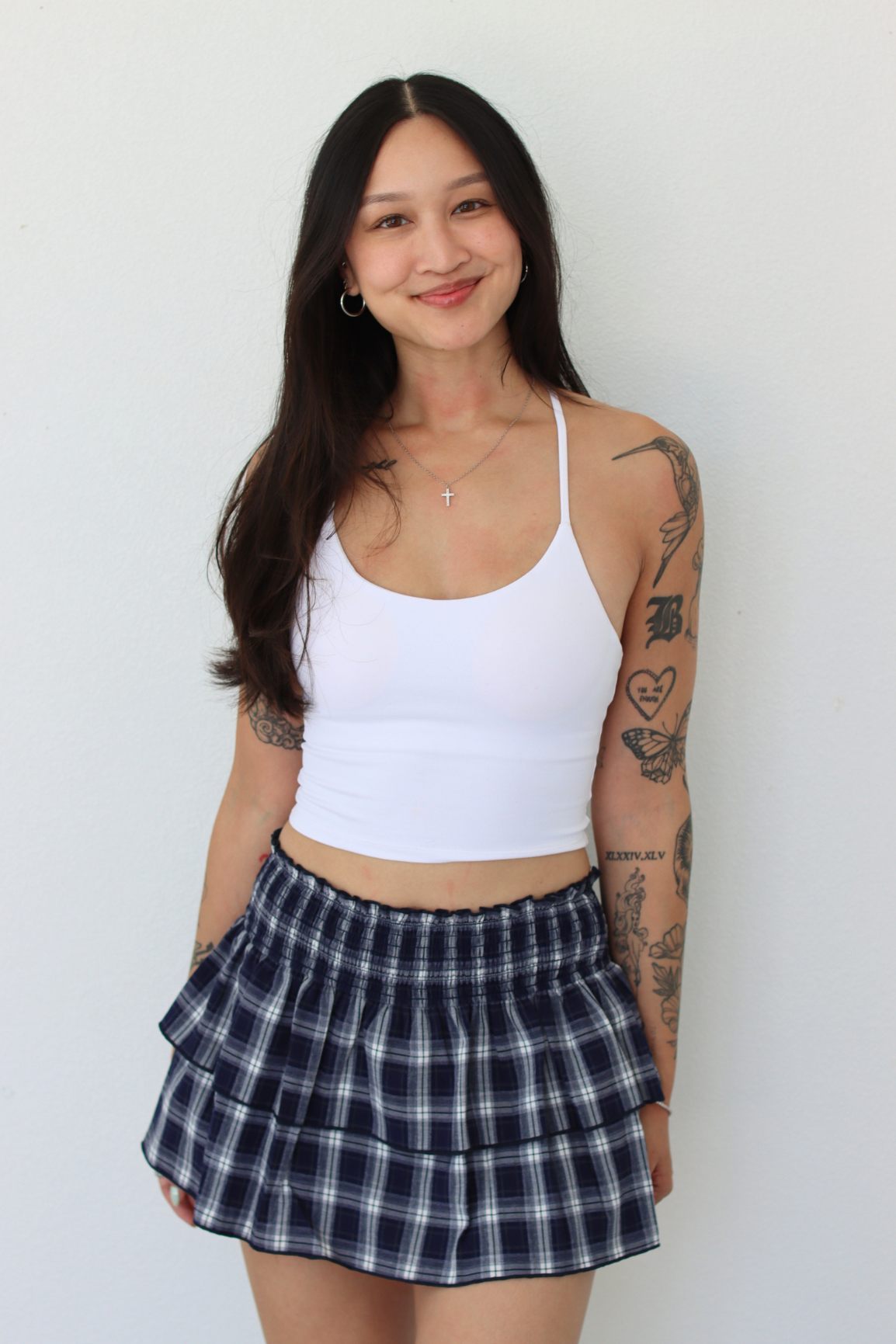 A woman with long dark hair and visible arm tattoos is wearing a white ribbed tank top and a navy and white plaid pleated mini skirt, standing against a plain off-white wall.
