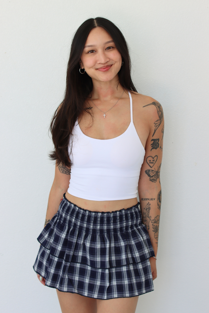 A woman with long dark hair and visible arm tattoos is wearing a white ribbed tank top and a navy and white plaid pleated mini skirt, standing against a plain off-white wall.