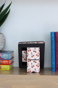 kindle with cherry pattern on a wooden surface with colorful books and a plant in the background.
