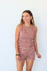 A woman modeling a sleeveless red and white gingham top with a smocked bodice and matching shorts, standing against a plain white background.