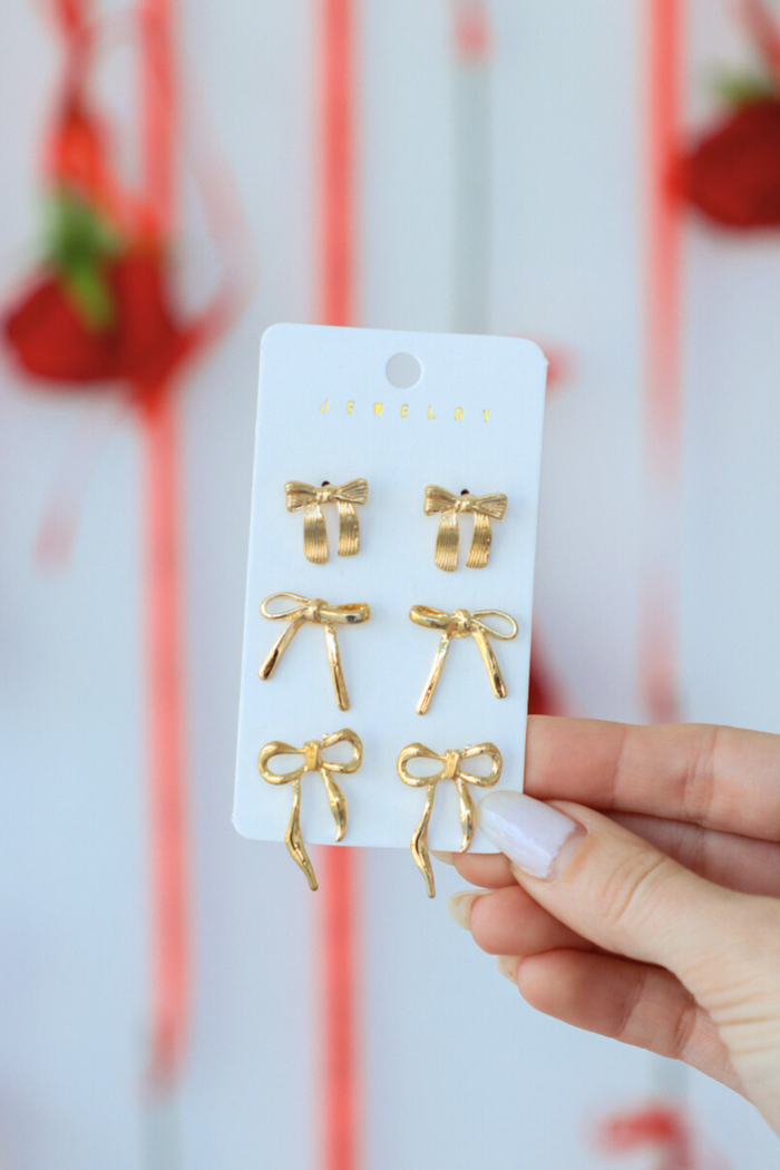 Gold bow-shaped earrings on a display card held by a hand with a blurred red and white background.