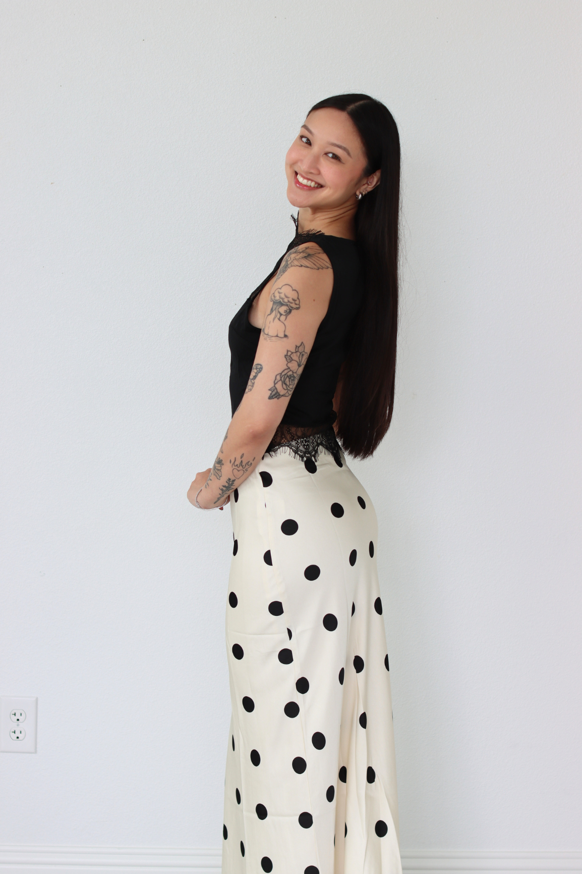 Woman wearing a black top and white polka dot skirt against a plain white wall.