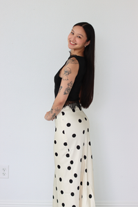 Woman wearing a black top and white polka dot skirt against a plain white wall.