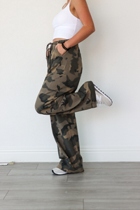 Person wearing camouflage pants and a white top, standing against a white wall.