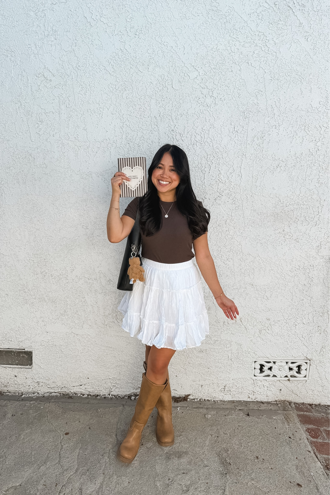 A woman posing in front of a white stucco wall, wearing a brown short-sleeve top, a white tiered ruffled mini skirt, and tan knee-high boots, while holding a striped kindle case.