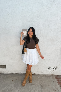 A woman posing in front of a white stucco wall, wearing a brown short-sleeve top, a white tiered ruffled mini skirt, and tan knee-high boots, while holding a striped kindle case.