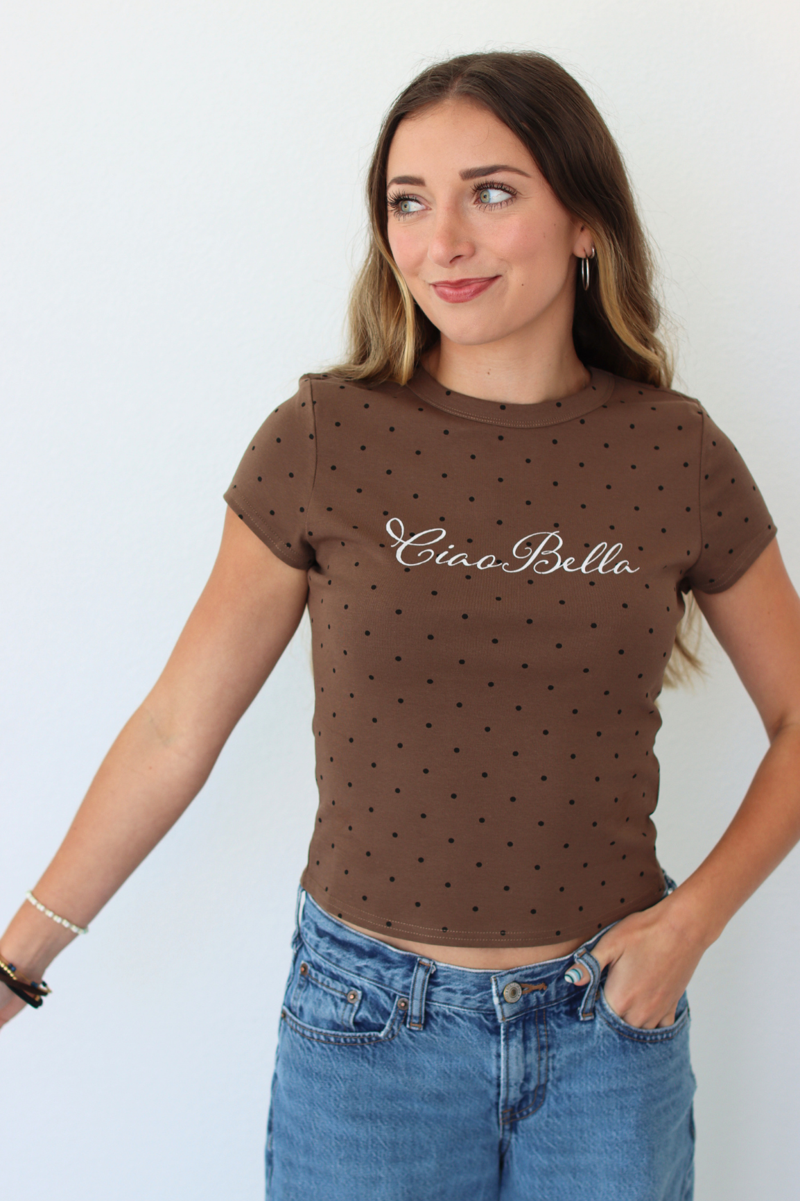A woman wearing a brown short-sleeve crop top with small black polka dots and the text 'Ciao Bella' in white script across the chest, paired with blue jeans, against a plain white background.