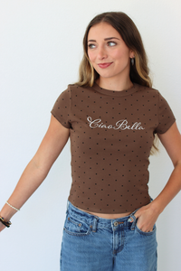 A woman wearing a brown short-sleeve crop top with small black polka dots and the text 'Ciao Bella' in white script across the chest, paired with blue jeans, against a plain white background.