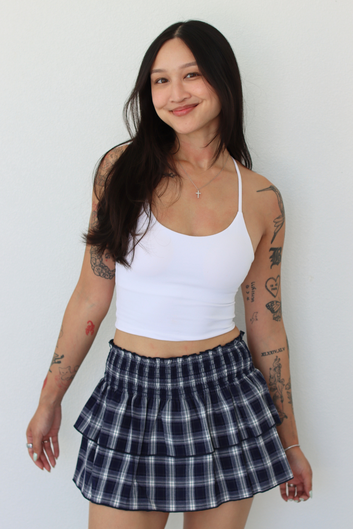 A woman with tattoos is wearing a white ribbed crop top and a navy blue and white plaid tiered pleated mini skirt, standing against a plain white wall.