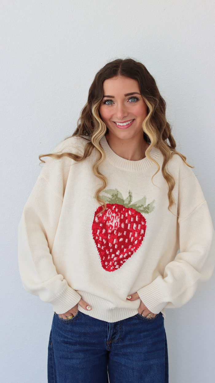 Woman wearing a cream sweater with a strawberry design against a white background