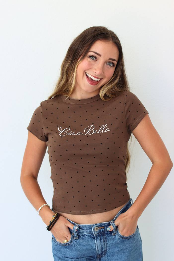 A woman smiling and wearing a brown short-sleeve crop top with small black polka dots and the text 'Ciao Bella' printed across the chest, paired with blue jeans against a plain white background.