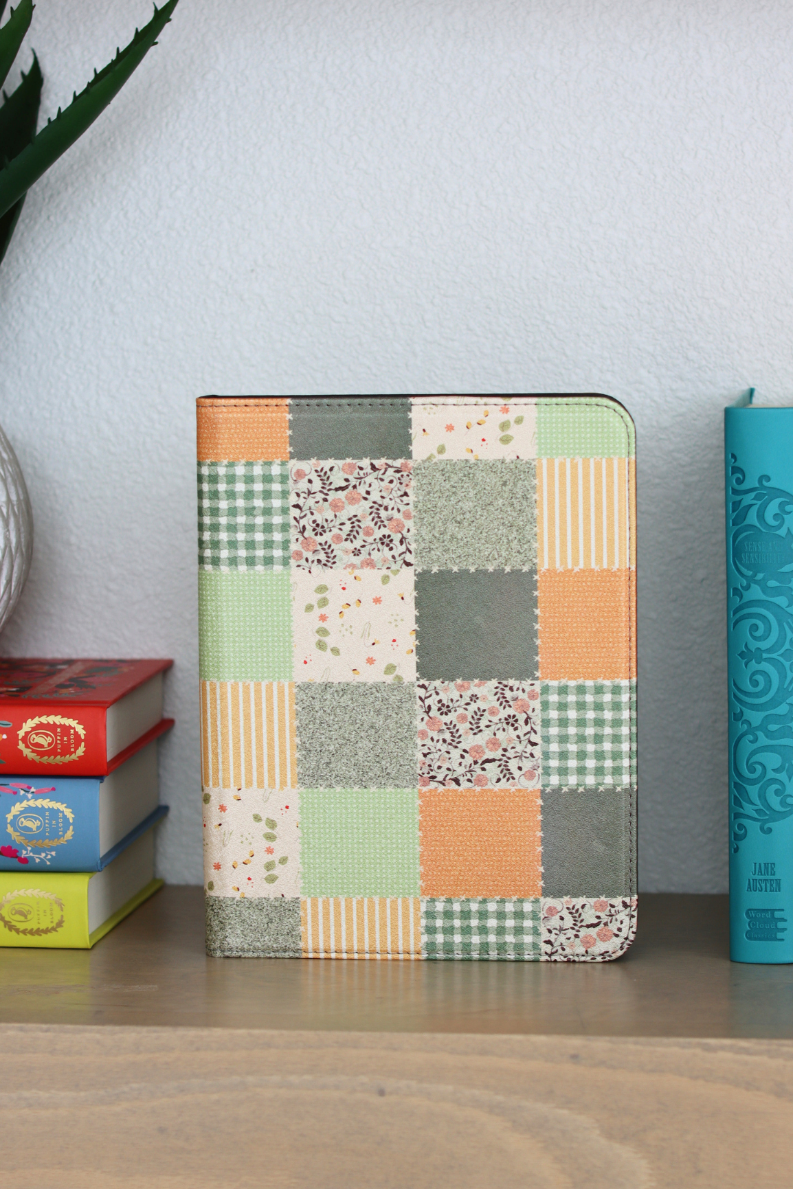 A nook case with a multicolor patchwork fabric design featuring orange, green, beige, and gray squares, standing on a wooden shelf next to stacked books.