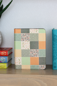 A nook case with a multicolor patchwork fabric design featuring orange, green, beige, and gray squares, standing on a wooden shelf next to stacked books.