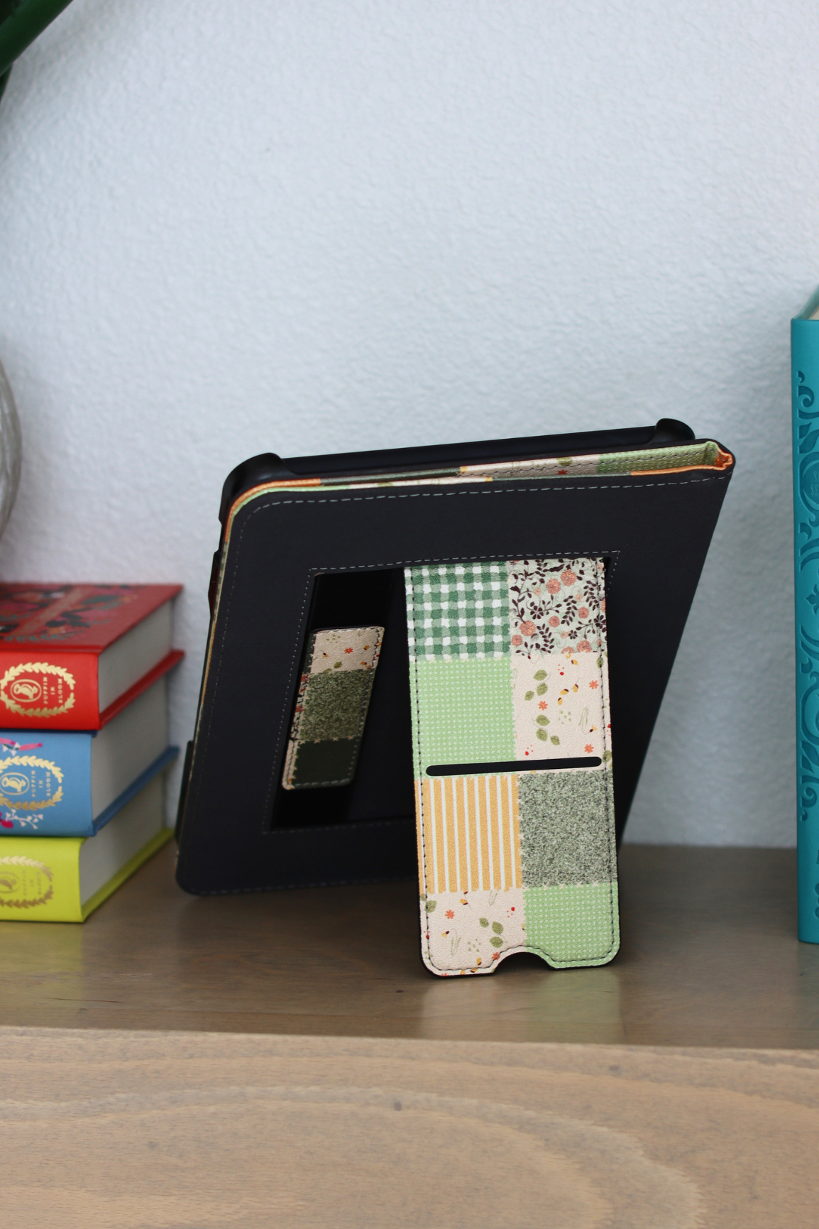 A nook case with a built-in kickstand and hand strap, displayed on a wooden surface next to books against a light blue wall.