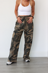 Person wearing camouflage pants and a white top on a plain background