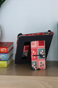 A pink nook case with a built-in stand, featuring a colorful floral patchwork pattern on the front cover, displayed on a wooden shelf next to a stack of books against a light blue wall.