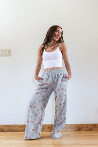 Woman wearing a white tank top and patterned gray pajama pants standing against a plain wall.
