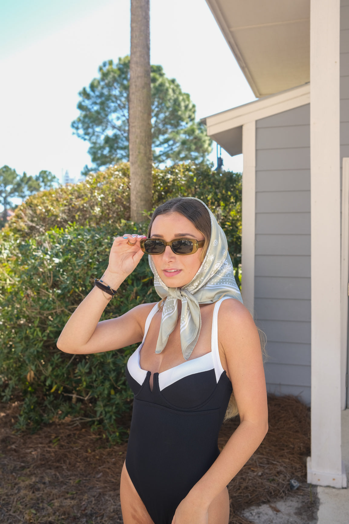 Woman wearing a black and white swimsuit with sunglasses and a headscarf outdoors.