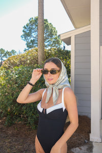Woman wearing a black and white swimsuit with sunglasses and a headscarf outdoors.