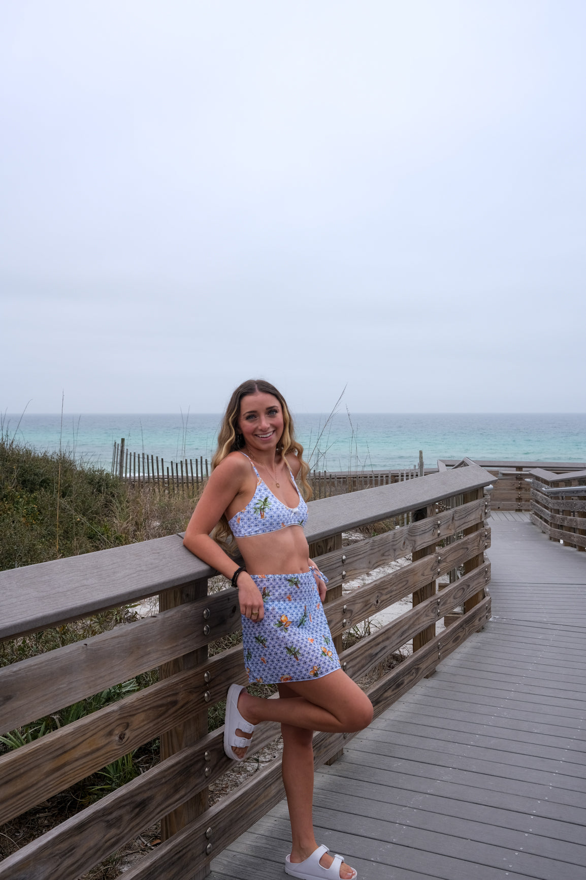 Woman in a patterned outfit standing on a wooden boardwalk by the ocean.