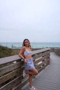 Woman in a patterned outfit standing on a wooden boardwalk by the ocean.