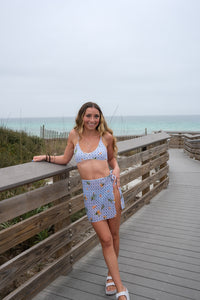 Woman in a blue patterned outfit standing on a wooden boardwalk by the beach.
