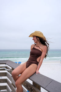 Woman in a brown swimsuit and straw hat sitting on a wooden bench by the beach.