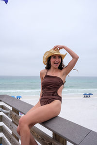 Woman in a brown swimsuit and straw hat sitting on a wooden bench at the beach.