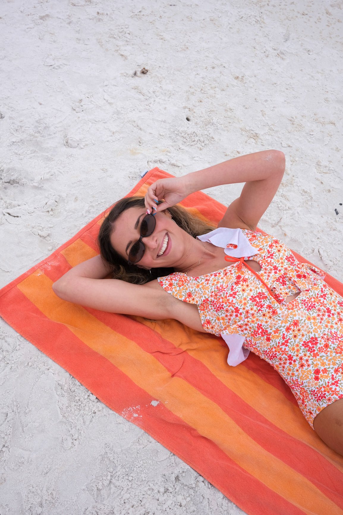 Woman in a floral dress lying on an orange striped towel at the beach.