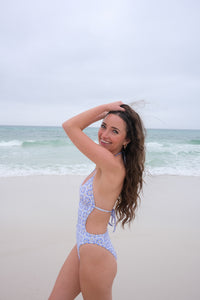 Woman in a light blue floral swimsuit standing on a beach with ocean waves in the background.